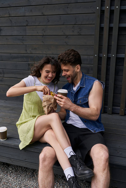 Pozitivní brunetka žena v botách a slunečních šatech drží čerstvou housku v blízkosti stylového přítele v džínové vestě s kávou jít, zatímco sedí v blízkosti dřevěného domu na pozadí, klidné prostředí koncepce - Fotografie, Obrázek