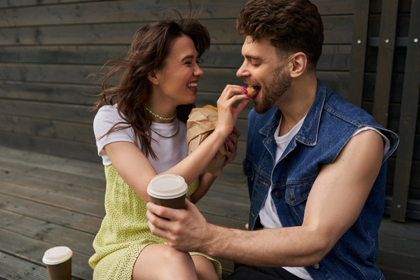 Femme brune gaie et élégante en robe de soleil nourrissant petit ami avec chignon et assis près du café pour aller et maison en bois à l'arrière-plan, concept d'ambiance sereine, tranquillité - Photo, image