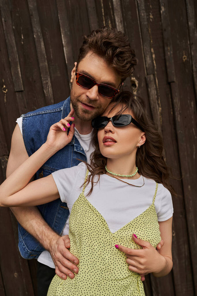 Portrait of stylish bearded man in sunglasses and denim vest embracing brunette girlfriend in sundress and standing together near wooden building outdoors, countryside exploration concept - Foto, afbeelding