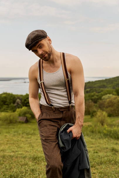 Портрет модного і бородатого чоловіка в шапці і вінтажному вбранні тримає руку в кишені і піджаку, стоячи з сільським пейзажем на фоні, мальовнича концепція сільської місцевості - Фото, зображення