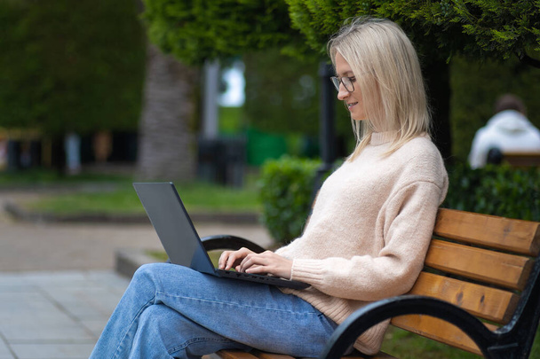 Smiling blonde young woman with glasses is working on a laptop, typing on a keyboard, sitting on a park bench. Side view of a student with glasses studying online outdoors. Freelancer in the park - Photo, Image