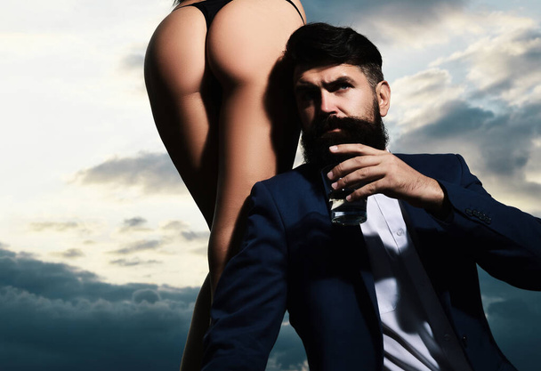 Rich man drink brandy. Bearded businessman in elegant suit with glass of whiskey. Passionate couple, sensual lovers. Woman in bikini - Zdjęcie, obraz