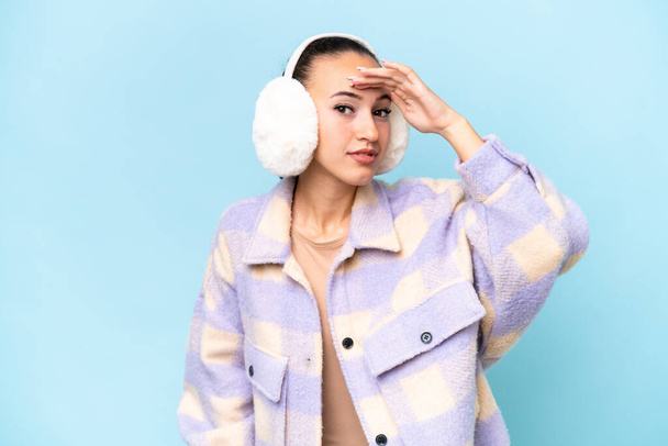 Young Arab woman wearing winter muffs isolated on blue background looking far away with hand to look something - Fotografie, Obrázek