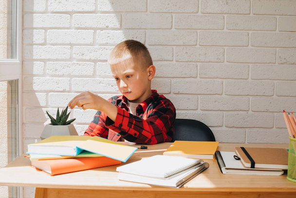 Wracajcie do szkoły. Chłopiec w trakcie nauki siedzi przy stole z książkami, notatnikami i ołówkami. Widok z przodu - Zdjęcie, obraz