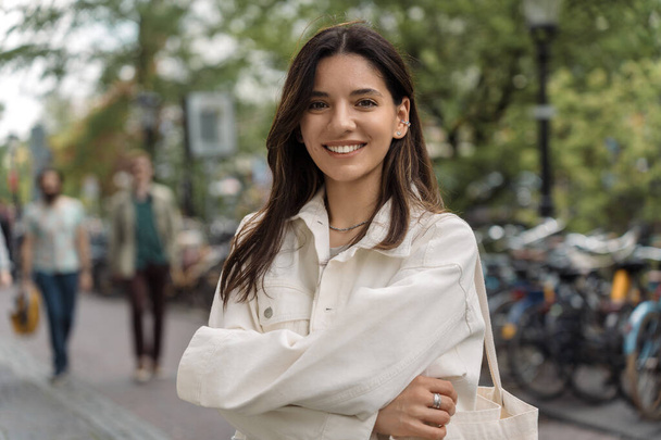 Portrait of multiethnic strong LGBT gorgeous woman confidently smiling hugging herself on the street of blurred city in background. - Фото, изображение