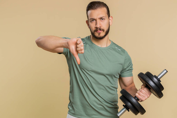 Upset tired athlete sportive man showing thumbs down sign gesture, expressing discontent, disapproval, dissatisfied, dislike. Middle eastern sportsman guy weightlifting dumbbell on beige background - Photo, Image