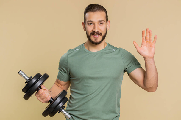 Come here, join us. Welcome. Athlete sportive man showing inviting gesture with hands, ask to join, beckoning to coming, gesturing hello or goodbye. Sportsman guy with dumbbell on beige background - Photo, Image