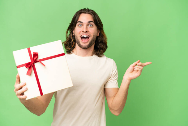 Young handsome man holding a gift over isolated background surprised and pointing finger to the side - Photo, image