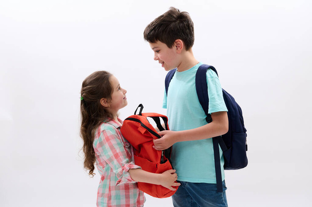 Caucasian happy children, preteen boy and preschooler little kid girl with backpacks, smile looking at each other, isolated over white studio background. Education. Back to school concept - Photo, Image