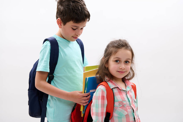 Ένα έφηβο αγόρι που βοηθάει τη μικρότερη αδερφή του να πακετάρει τη σχολική της τσάντα, απομονωμένη σε λευκό φόντο στούντιο. Φιλίες. Βοήθεια και υποστήριξη. Αδελφός και αδελφή πηγαίνουν στο σχολείο - Φωτογραφία, εικόνα