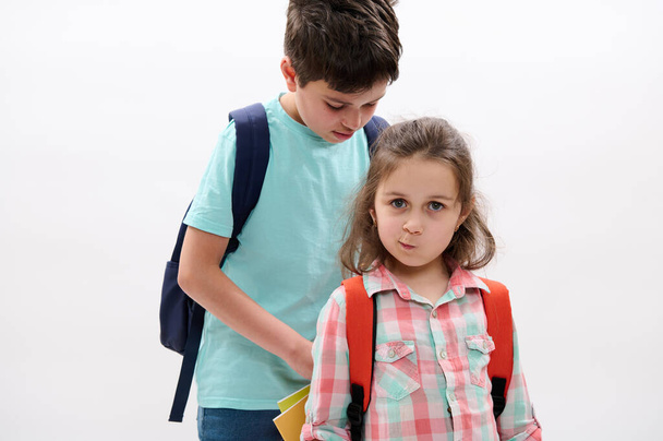 Lovely little child girl, mischievous preschooler kid makes faces looking at camera while her older brother puts workbooks inside her backpack, isolated over white background. Back to school concept - Foto, imagen
