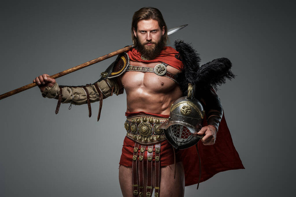 Parrakas gladiaattori, jolla on pitkät hiukset, seisoo harmaalla pohjalla kevyessä haarniskassa ja punaisessa kaavussa, pitäen keihästä ja kypärää höyhenillä. - Valokuva, kuva