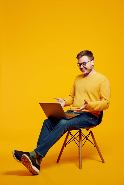 Young confused man wearing yellow sweater and eyeglasses, using laptop while sitting on chair, isolated over orange background - Zdjęcie, obraz