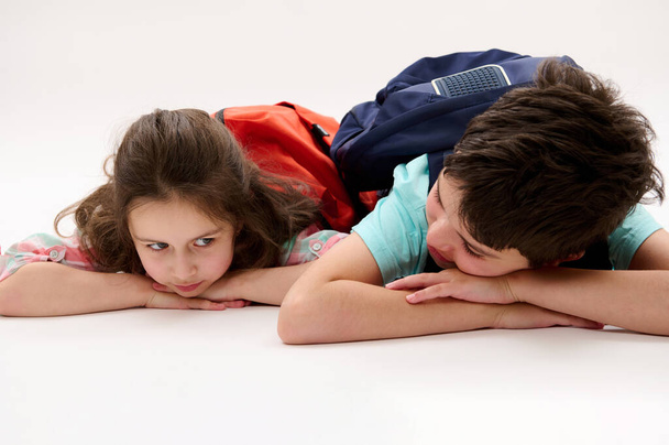 Conceptual emotional portrait of a happy little preschool girl and her brother, preteen school boy with backpacks, talking to each other in school recreation time, lying on belly over white background - Фото, зображення