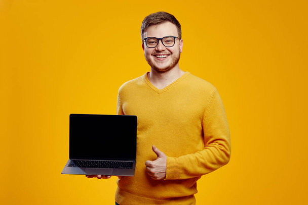 Izgalmas szakállas férfi sárga pulóvert és szemüveget visel, gesztusként viselkedik, miközben új, modern laptopot tart, narancssárga háttér felett elszigetelve. - Fotó, kép