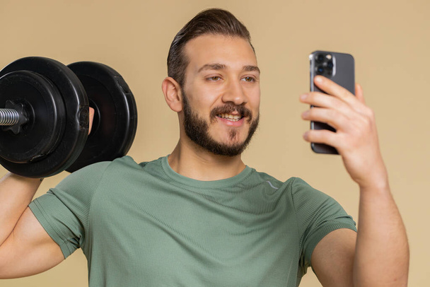 Athlete sportive man texting share messages content on smartphone social media applications online, sms message, browsing, reading news. Middle eastern sportsman guy with dumbbell on beige background - Foto, Bild
