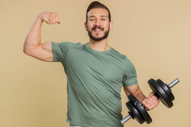 I am strong. Athlete sportive man showing biceps and looking confident, feeling power strength, energy to gain success win, motivation. Middle eastern sportsman guy with dumbbell on beige background - Valokuva, kuva