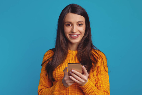 Młoda atrakcyjna dziewczyna z dużym uśmiechem, trzymając smartfona, cieszy się komunikacją online, odizolowany na niebieskim tle - Zdjęcie, obraz