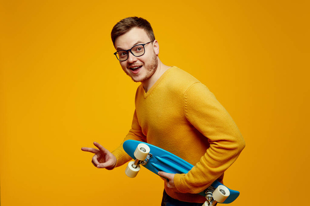 Happy young man in yellow sweater and eyeglasses holding blue longboard and showing v sign gesture against orange background - Photo, Image