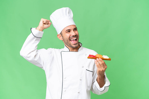 Joven chef caucásico sosteniendo un sushi sobre un fondo aislado celebrando una victoria - Foto, imagen