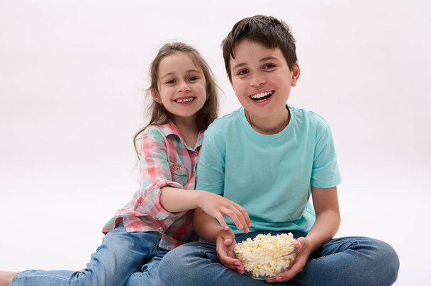 Caucasian happy cute children, preteen boy and little girl, brother and sister sitting over white isolated studio background, watching cartoons or movie, eating popcorn. kids. Lifestyle. Entertainment - Foto, afbeelding