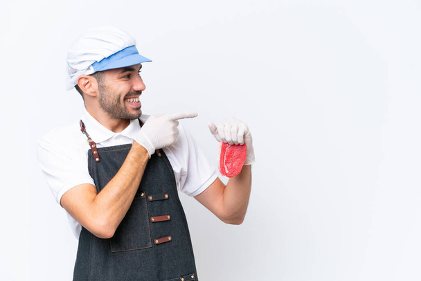 Butcher caucasian man wearing an apron and serving fresh cut meat over isolated white background pointing finger to the side and presenting a product - Φωτογραφία, εικόνα