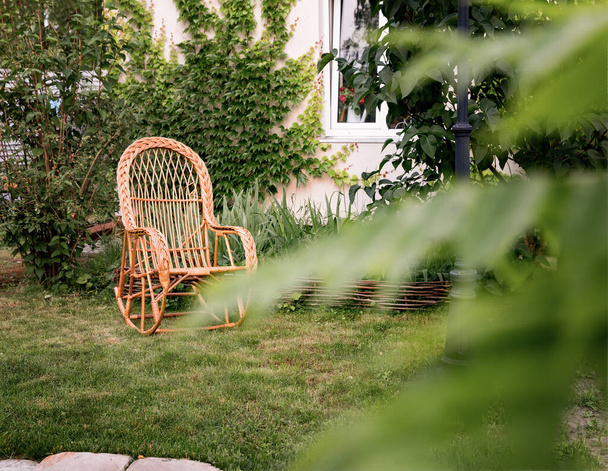 Een grote mooie rieten stoel in de tuin tegen de achtergrond van het huis. Veel struiken en vegetatie. Naast de stoel is een kleine bloementuin. - Foto, afbeelding
