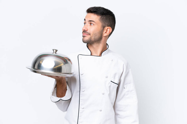 Junger Koch mit Tablett auf weißem Hintergrund, der zur Seite schaut und lächelt - Foto, Bild