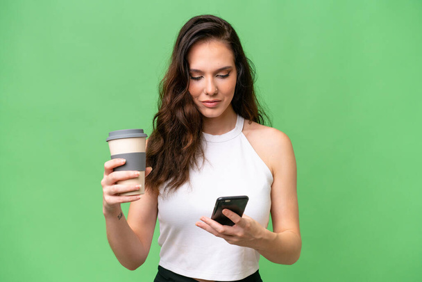 Junge kaukasische Frau isoliert über isolierten Hintergrund mit Kaffee zum Mitnehmen und ein Handy - Foto, Bild