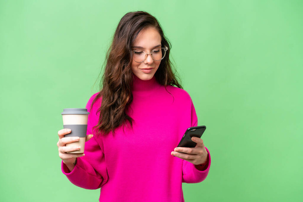 Junge kaukasische Frau isoliert über isolierten Hintergrund mit Kaffee zum Mitnehmen und ein Handy - Foto, Bild