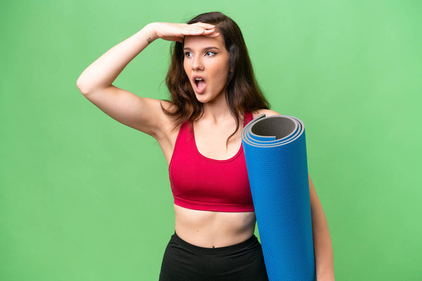 Young sport woman going to yoga classes while holding a mat over isolated background doing surprise gesture while looking to the side - Photo, Image