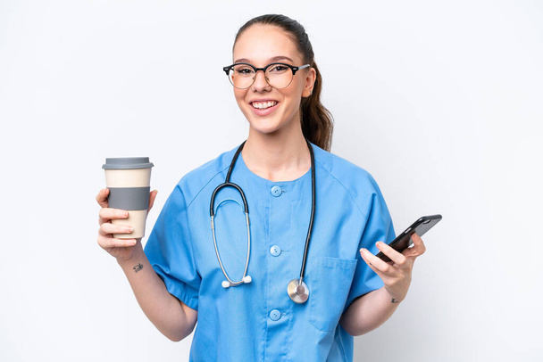 Young caucasian surgeon doctor woman isolated on white background holding coffee to take away and a mobile - Φωτογραφία, εικόνα