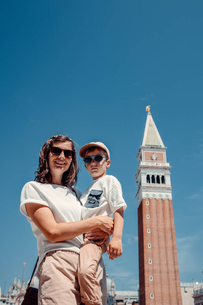 Μια μητέρα και το τετράχρονο αγόρι της αιχμαλωτίστηκαν σε μια εγκάρδια στιγμή, φωτογραφήθηκαν στην κεντρική πλατεία της Βενετίας. Ο δεσμός τους ακτινοβολεί μέσα από το φακό, ενσωματώνοντας την ουσία της αγάπης και της οικογένειας μέσα στην ομορφιά της εμβληματικής πόλης της Ιταλίας. - Φωτογραφία, εικόνα