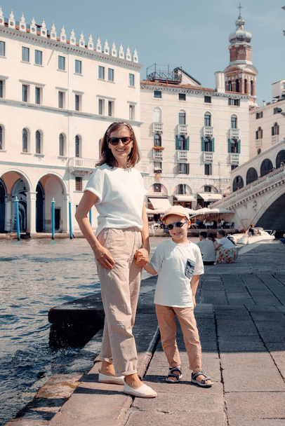 Η αγνή χαρά ακτινοβολεί από ένα όμορφο παιδί καθώς περπατούν μαζί με τη μητέρα τους στους μαγευτικούς δρόμους της Βενετίας. - Φωτογραφία, εικόνα