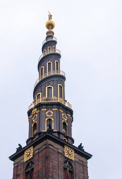 Copenhagen, Denmark - September 14, 2010: Vor Frelsers church. Tower closeup against light blue sky. Golden trim of spiral and clock. Statues on corniche and on golden globe at top - Photo, Image