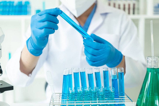 Ένας επιστήμονας γάντια ρίχνει μπλε οξύ σε ένα δοκιμαστικό σωλήνα με διαυγές υγρό. Επιστημονική έρευνα στη χημική βιομηχανία Ο γιατρός εξετάζει την ανάλυση σε δοκιμαστικό σωλήνα με μικροσκόπιο. - Φωτογραφία, εικόνα