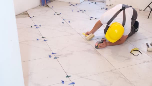 Professional laying of ceramic tiles on a concrete floor. The worker is dressed in overalls and a construction helmet, making repairs in the house - Materiaali, video
