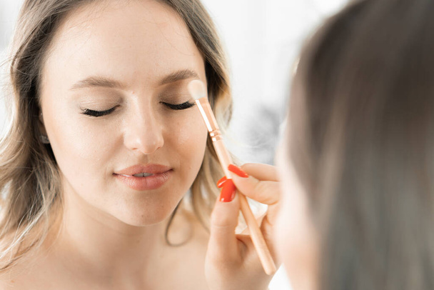De processen van make-up van een jonge vrouw in een schoonheidssalon vinden plaats: de make-up kunstenaar brengt vakkundig een dunne laag van de basis aan op haar gezichtshuid, op de oogleden, voor verdere make-up. Bruiloft - Foto, afbeelding