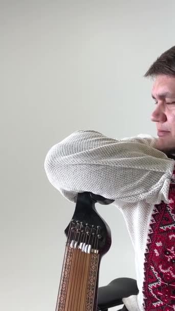 fuerte cosaco ucraniano en una camisa bordada punto de cruz roja en un lino blanco apoyado en una bandura instrumento musical y mira a la distancia  - Imágenes, Vídeo