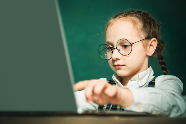 Pupil working on laptop computer over blackboard background. Child near chalkboard in school classroom. Kid is learning in class on background of blackboard - 写真・画像