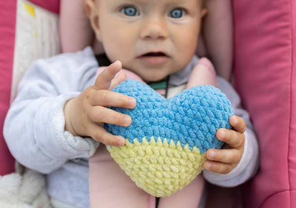 Un petit bébé innocent tient un cœur ukrainien jaune-bleu dans ses mains. Les enfants sont priés d'arrêter la guerre en Ukraine. Réfugiés amoureux de l'Ukraine. Jour de l'indépendance - Photo, image
