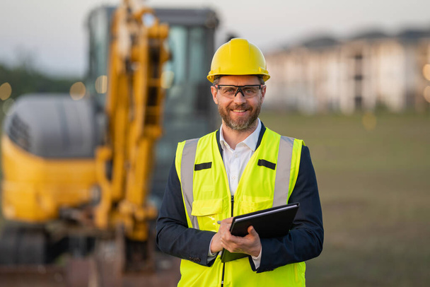 Architect at a construction site. Architect man in helmet and suit at modern home building construction. Architect with a safety vest and suit. Confident architect standing at house background - Foto, Bild