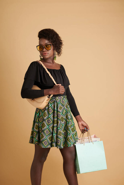 Elegante confiante preto jovem mulher em óculos de sol carregando sacos de compras e olhando para longe - Foto, Imagem