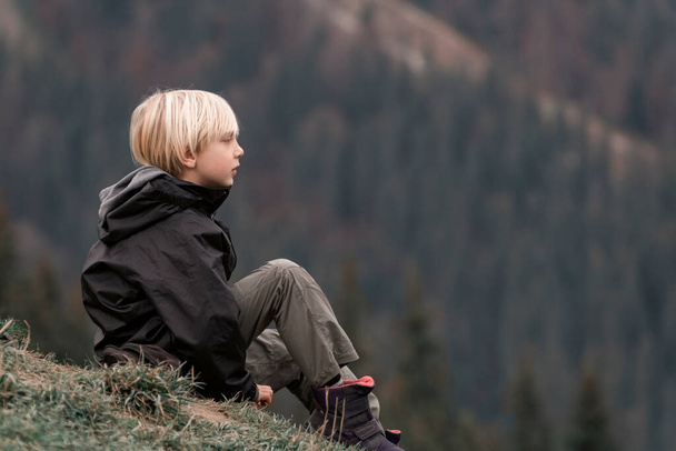 Child on the mountainside. Sad brooding blond boy in trekking clothes on hike during break - Zdjęcie, obraz