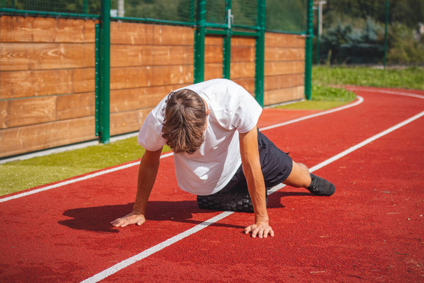 Ein braunhaariger Junge mit athletischem Körperbau auf einem athletischen Oval massiert seine Muskeln mit einer Schaumstoffrolle, um sich besser zu erholen. Post-workout. - Foto, Bild