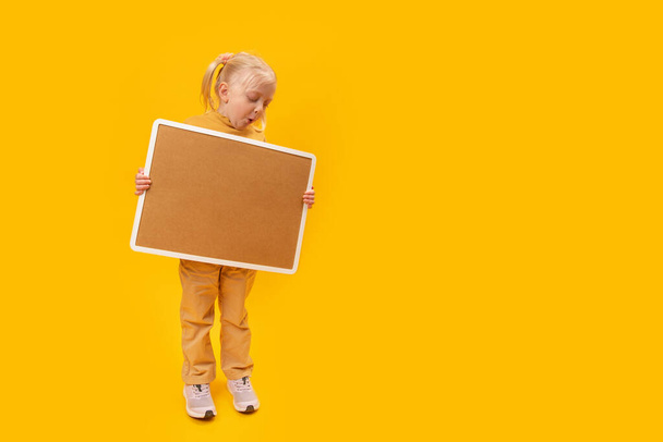 Full length portrait of cute caucasian child girl on yellow background with cork board holding in hand. Copy space, mockup. - Valokuva, kuva
