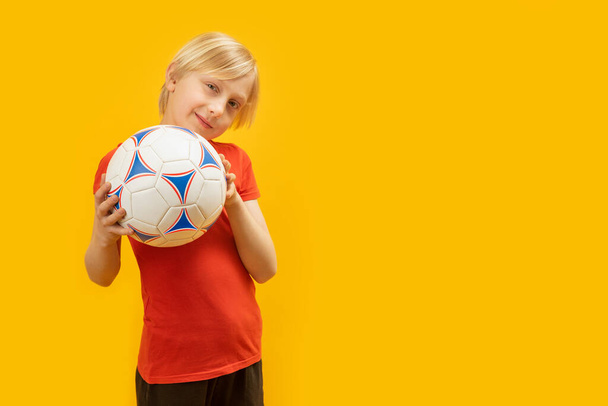 Blonďák v červeném tričku drží fotbalový míč. Studio portrét teenagera s míčem na žlutém pozadí. Kopírovat prostor - Fotografie, Obrázek