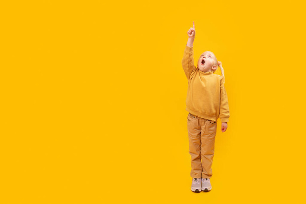 驚いた少女は指を上に向けている。明るい黄色の背景に就学前の女の子の完全な長さの肖像画。コピースペース,テキストのためのスペース. - 写真・画像