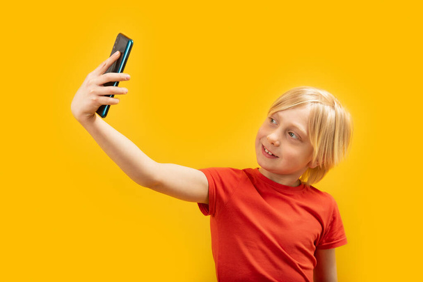 Portrait of blond boy in red T-shirt with phone in his hands. Child takes selfie. Isolated on yellow background. - Fotó, kép