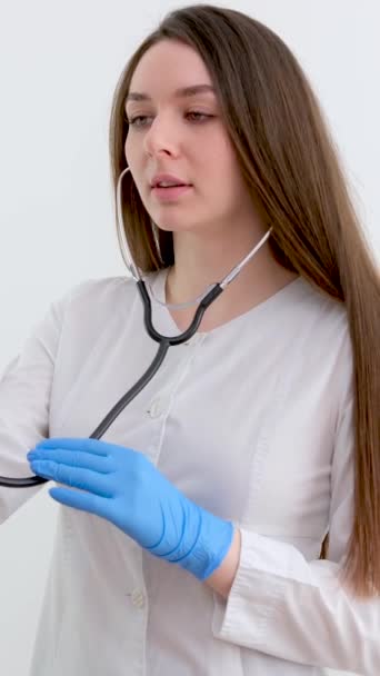 Nuori naispuolinen kardiologi valkoisessa takissa fonendoskoopin avulla kuuntelee nuoren naisen sykettä klinikan toimistossa tehtävän tarkastuksen aikana. Laadukas 4k kuvamateriaalia - Materiaali, video